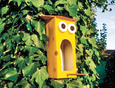 Gelber bemalter Getränkekarton mit Gesicht hängt vor einer grünen Hecke.